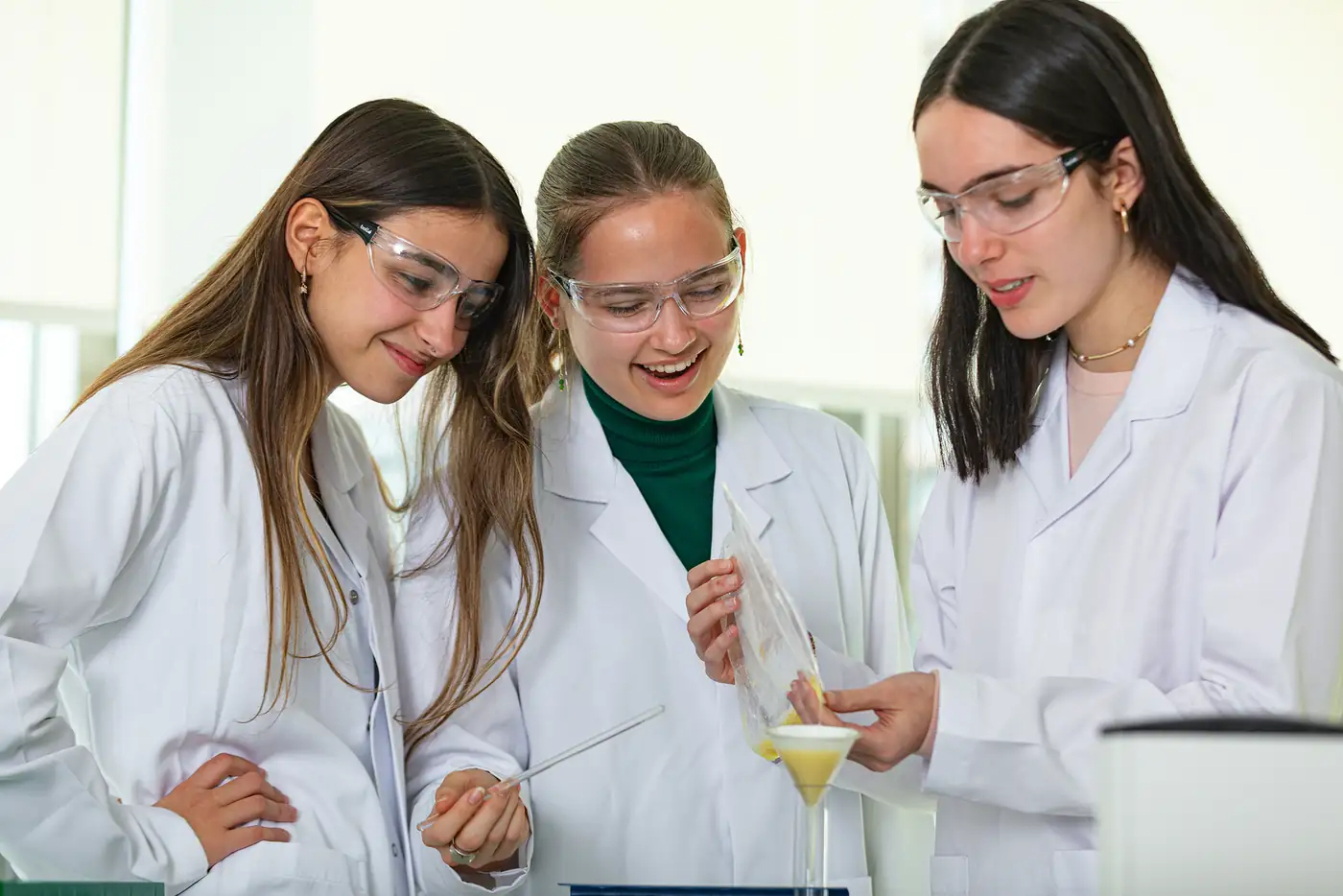 Three senior students in lab coats and safety glasses carry out a chemistry experiment